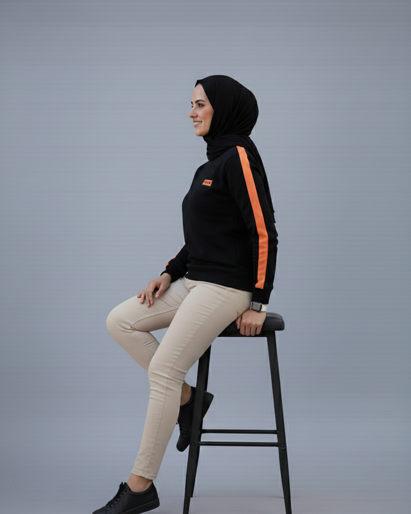 Woman wearing a black hoodie with orange stripes and beige pants sitting on a stool against a gray background