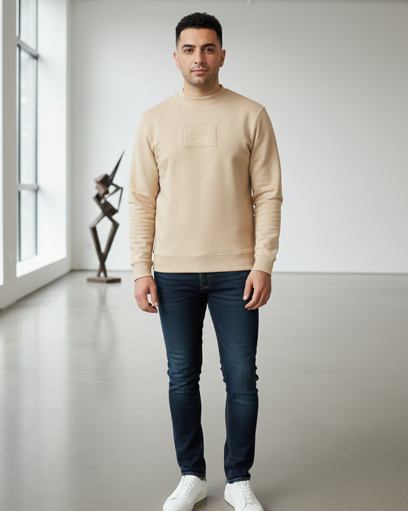 Man wearing a beige sweater, dark jeans, and white shoes in a modern indoor setting.