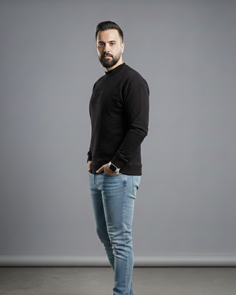 Man wearing a black sweater and light blue jeans standing against a gray background